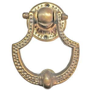 May include: A brass door knocker with a rounded, ornate design. The knocker has a textured surface and a ring-shaped handle.