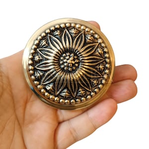 May include: A circular, antique brass-toned decorative piece featuring a sunflower motif. The sunflower is centrally positioned, encircled by a beaded edge. The object is held in a hand against a white backdrop.