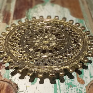 May include: A circular, brass-toned decorative item with an elaborate openwork pattern. The central design includes a floral motif, encircled by concentric rings and a scalloped border. This piece is likely intended for decorative purposes.