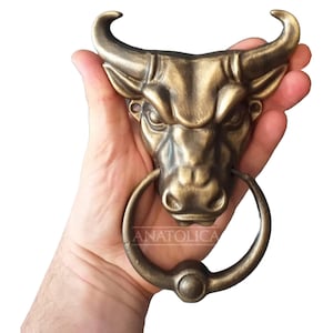 May include: A gold bull's head door knocker with a circular ring. The knocker is held in a hand.