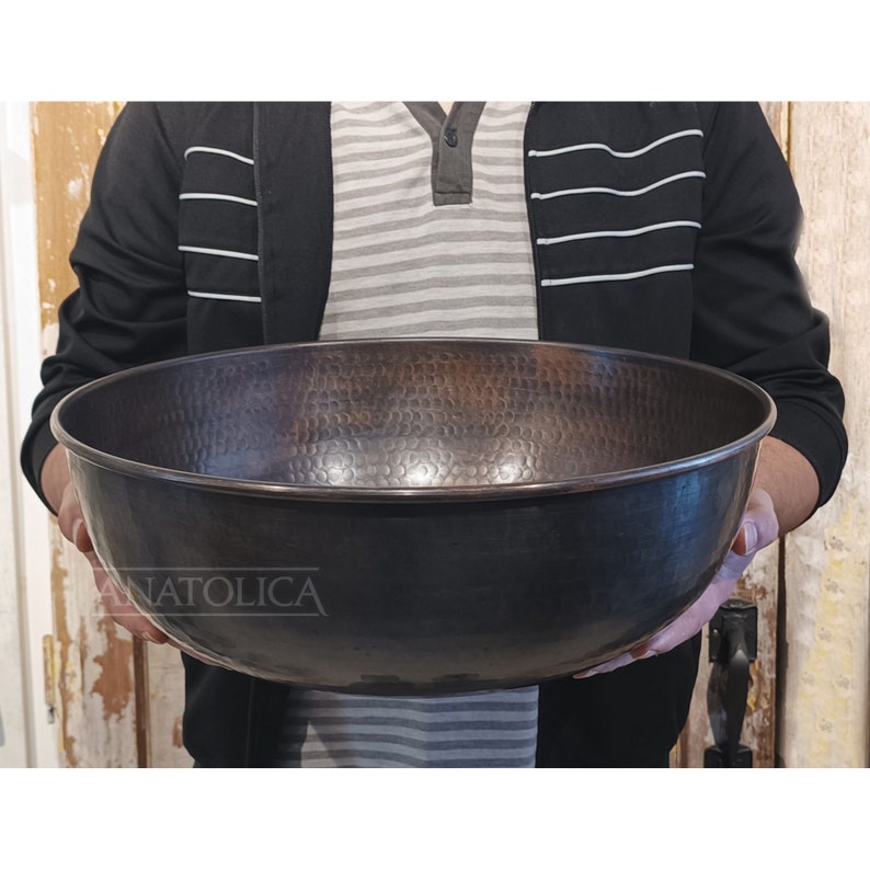 May include: A large, hammered copper bowl with a dark patina finish. The bowl is being held by a person's hands.