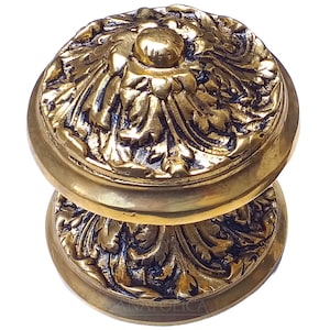 May include: Ornate brass door knob with a detailed floral design. The knob is a warm gold colour and features a raised central element. The design is symmetrical and the knob appears to be vintage or antique.