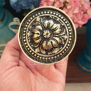 May include: A round, gold-toned metal knob with a decorative floral design. The knob has a raised, ornate border.