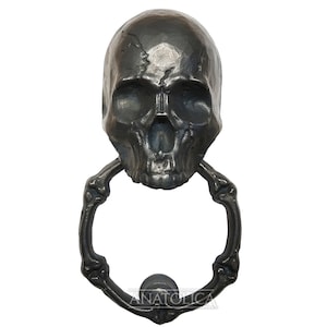 May include: Black metal skull door knocker with a bone-shaped ring. The skull has a detailed design with a cracked surface.