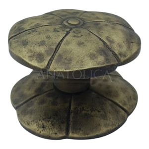 May include: A bronze-colored cabinet knob with a floral design. The knob has a rounded top and base, with a central stem. The surface has a textured, aged appearance. The word "ANATOLICA" is printed on the base.