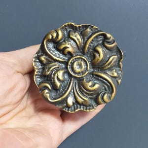 May include: A round, ornate brass cabinet knob with a floral design. The knob has a dark patina and a raised centre.