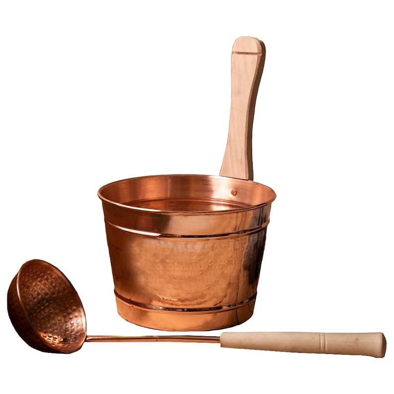 May include: A copper sauna bucket with a wooden handle and a matching copper ladle with a wooden handle. The bucket has a hammered finish.