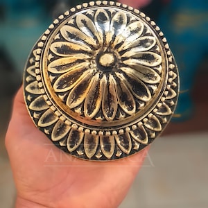 May include: A gold-toned metal door knob with a decorative floral design. The knob has a raised, circular centre with a smaller, circular design in the centre. The knob is surrounded by a ring of smaller, decorative flowers.