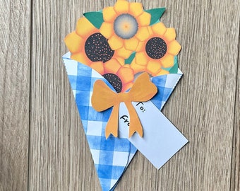 Può includere: Un cono di carta a quadri blu e bianchi pieno di girasoli gialli con centri neri. Un fiocco giallo è attaccato al cono. Un biglietto bianco con la parola "A:" scritta su di esso è attaccato al cono.