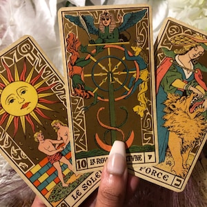 Puede incluir: Tres cartas de tarot, el Sol, la Rueda de la Fortuna y la Fuerza, con una mano sosteniendo las cartas. Las cartas están ilustradas con imágenes tradicionales del tarot, incluyendo un sol, una rueda y una mujer con un león.