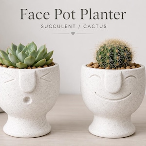 Può includere: Due vasi in ceramica bianca maculata a forma di volto con base a piedistallo. Il vaso a sinistra presenta un'espressione sorpresa con una piccola bocca rotonda e contiene una succulenta verde. Il vaso a destra ha un volto sorridente e un cactus.