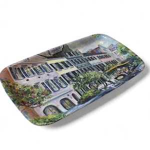 May include: A rectangular serving tray featuring a colorful painted scene of a street lined with multi-story buildings, dark shutters, arched doorways, and lush green trees. The artwork depicts a city landscape with a horse-drawn carriage.