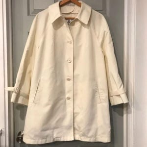 May include: Cream-colored trench coat with a classic design. Features a collared neckline, button-down front, and long sleeves with adjustable cuffs. Includes two front pockets and a mid-length hem. The coat is hanging on a wooden hanger.