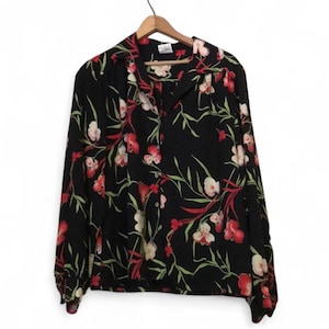 Könnte beinhalten: Schwarze Bluse mit floralem Muster aus roten und weißen Blüten und grünen Blättern. Das langärmlige Hemd hängt an einem Holzkleiderbügel.