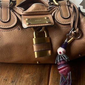 May include: A tan leather handbag featuring a gold-toned metal padlock closure with the brand name Chloe engraved on the plate. A small, felted clown-like charm with a purple hat, red nose, and multi-coloured fringed tassels hangs from the side ring.