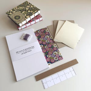 May include: A collection of mixed media paper and supplies. Includes a stack of white paper, patterned paper, a spool of purple thread, and a small booklet with floral designs. The text on the paper reads "48lb Canson XL Mixed Media Paper 8 inch x 5 inch Hand Trimmed Paper".