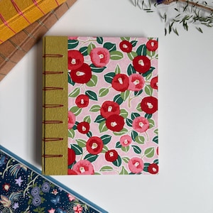 May include: A rectangular journal with a pink cover featuring a repeating pattern of red and pink camellia flowers with green leaves. The spine is covered in textured olive green paper and is bound with visible red thread stitching along the edge.