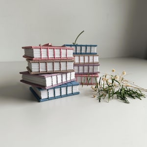 May include: A collection of handmade journals in various colors, including pink, blue, and purple. The journals are stacked and feature white pages and decorative stitching along the spine. A sprig of dried flowers adds a natural touch.