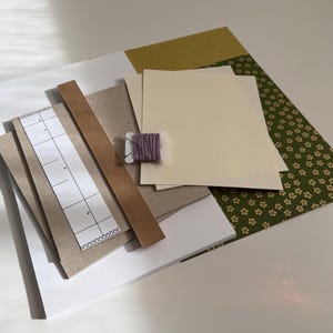May include: A craft supply kit featuring stacked rectangular sheets of white, cream, and brown cardstock, a green patterned paper with small floral motifs, a narrow brown wooden strip, a white paper template with grid markings, and a small spool of lavender thread.