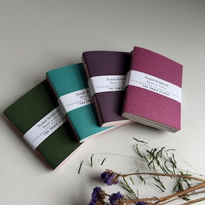 May include: Four colorful pocket notebooks in shades of green, teal, purple, and magenta. Each notebook is wrapped with a white band that includes text. The notebooks are stacked at an angle, with a few dried flowers in the foreground.