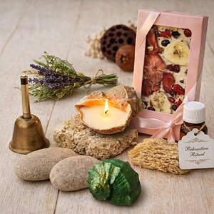 May include: A brass bell, a seashell candle, two smooth stones, a green seashell, a bundle of dried lavender, a lotus seed pod, a small brown fruit, and a pink gift box containing a white chocolate bar with dried fruit, next to a small oil bottle.
