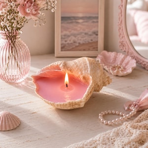 May include: A seashell candle featuring a natural tan exterior and a smooth, vibrant pink wax interior with a single lit wick. The shell is positioned horizontally, showcasing the contrast between the textured outer surface and the melted wax.