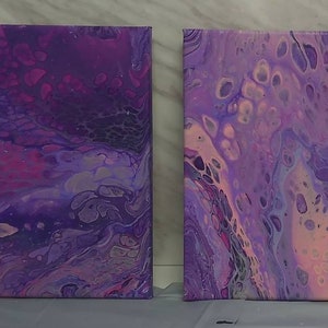 Op de afbeelding: Twee vierkante canvasdoeken met abstracte vloeibare kunstwerken in tinten paars, roze en wit. De schilderijen hebben een draaiende, marmerachtige uitstraling.