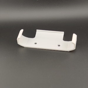 May include: A white, 3D-printed wall mount with two curved arms and two small holes. The mount is designed to hold an item, likely a small electronic device or accessory. The object is made of plastic.
