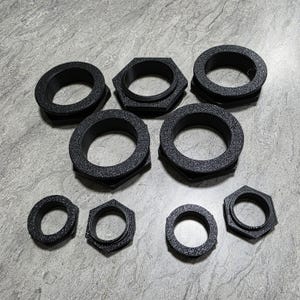 May include: A collection of black, textured, circular and hexagonal objects. The objects have a ring-like appearance with a central opening. The sizes vary, with some being larger and others smaller. The objects are arranged on a light gray surface.