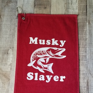 以下が含まれることがあります： 白いマスキーフィッシュのグラフィックと「Musky Slayer」というテキストが書かれた赤いゴルフタオル。