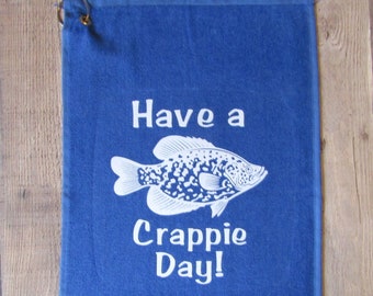 May include: A blue towel with the text "Have a Crappie Day!" and a white fish illustration. The towel has a metal hook for hanging.