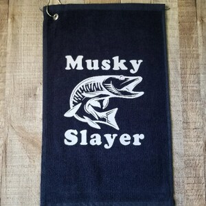 以下が含まれることがあります： 白いマスキーの魚のグラフィックと「Musky Slayer」というテキストが書かれた黒いゴルフタオル。