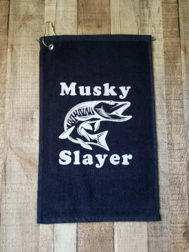 以下が含まれることがあります： 白いマスキーの魚のグラフィックと「Musky Slayer」というテキストが書かれた黒いゴルフタオル。
