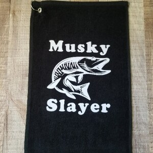 以下が含まれることがあります： 白いマスキーの魚のグラフィックと「Musky Slayer」というテキストが書かれた黒いゴルフタオル。