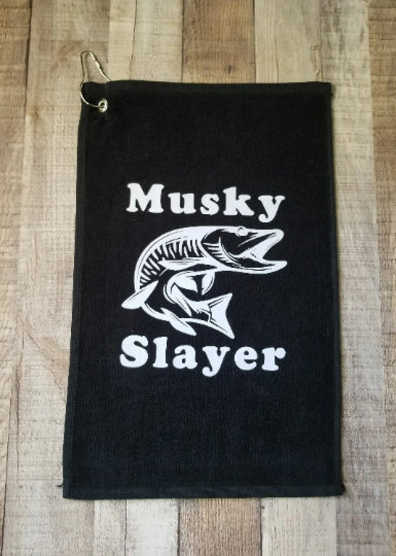 以下が含まれることがあります： 白いマスキーの魚のグラフィックと「Musky Slayer」というテキストが書かれた黒いゴルフタオル。