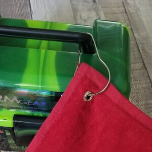 以下が含まれることがあります： 緑と黒のタックルボックスに金属製のリングが付いた赤いゴルフタオル。タオルの角には金属製のリングが付いており、タックルボックスにぶら下がっています。