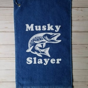 以下が含まれることがあります： 白いマスキーの魚のグラフィックと「Musky Slayer」というテキストが書かれた青いゴルフタオル。