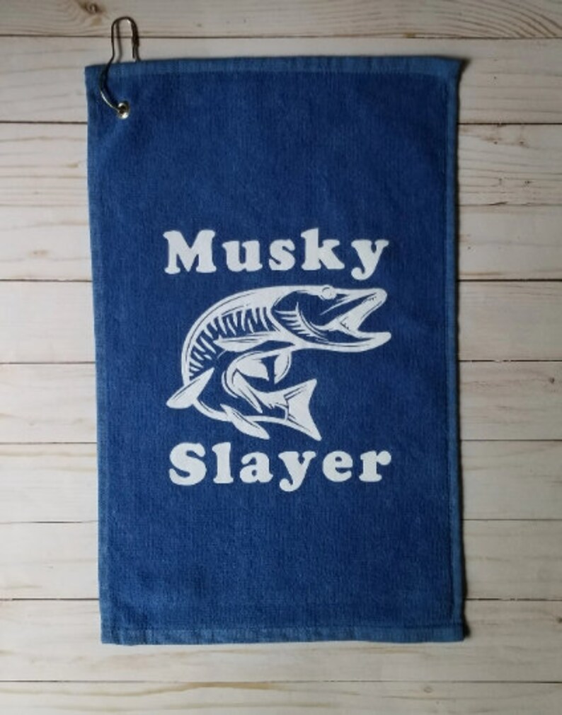 以下が含まれることがあります： 白いマスキーの魚のグラフィックと「Musky Slayer」というテキストが書かれた青いゴルフタオル。