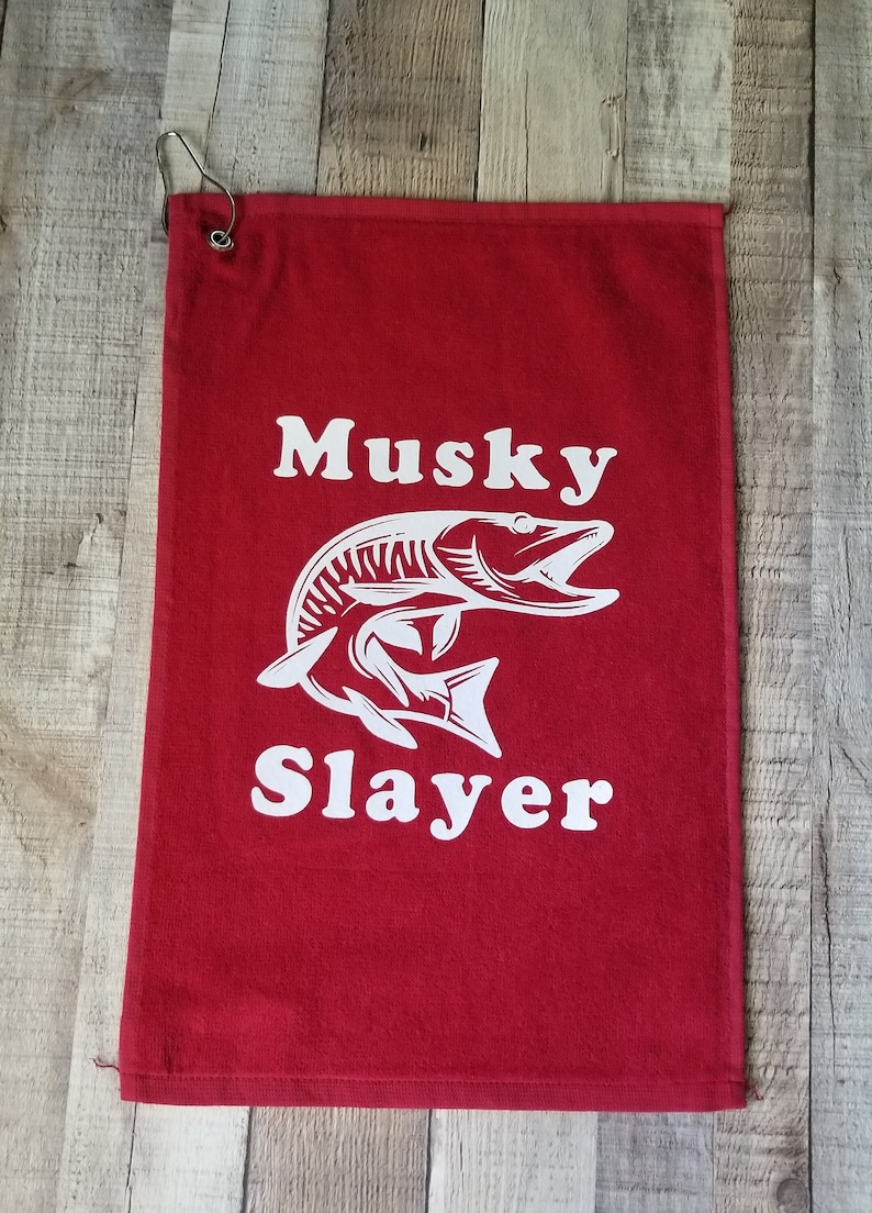 以下が含まれることがあります： 白いマスキーフィッシュのグラフィックと「Musky Slayer」というテキストが書かれた赤いゴルフタオル。