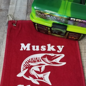以下が含まれることがあります： 赤い釣りタオルに、白いマスキーの魚のグラフィックと「Musky Slayer」の文字が書かれています。背景には、緑と黒のタックルボックスがあり、「Wormgear」の文字が書かれています。