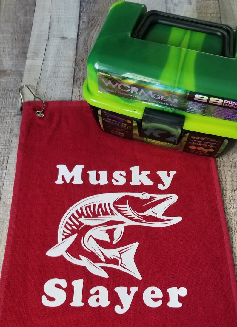以下が含まれることがあります： 赤い釣りタオルに、白いマスキーの魚のグラフィックと「Musky Slayer」の文字が書かれています。背景には、緑と黒のタックルボックスがあり、「Wormgear」の文字が書かれています。