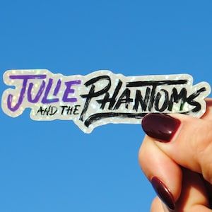 Puede incluir: Una pegatina blanca con acabado holográfico que muestra el texto "Julie and the Phantoms" en letras negras y moradas.