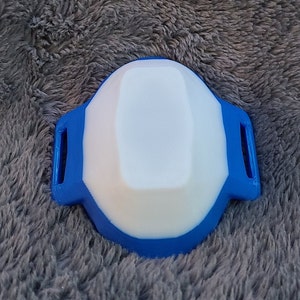 May include: A blue and white plastic device with a rounded shape and two small loops on the sides.