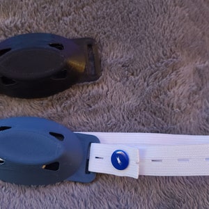 May include: Two 3D printed plastic holders for a medical device, one black and one blue, with a white elastic strap with a blue button.