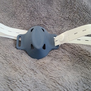May include: A gray 3D printed face mask with a white elastic strap. The strap has adjustable snaps.