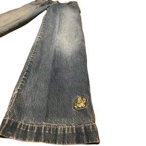 May include: A pair of blue denim jeans featuring a faded wash and a wide-leg silhouette. The lower portion of the left pant leg displays a small, embroidered patch of a stylized yellow-haired character face with dark outlines and expressive eyes.