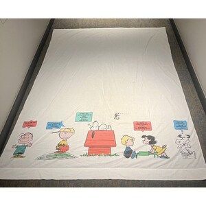 May include: White bed sheet featuring a colorful illustration of Snoopy and the Peanuts gang. The design includes Snoopy's doghouse, Charlie Brown, Lucy, and other characters with speech bubbles. The sheet has a cartoon style.
