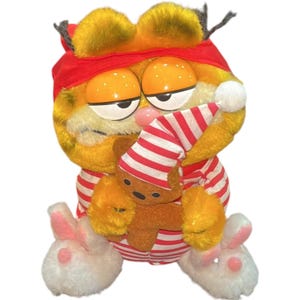 May include: A plush Garfield toy wearing a red sleep mask and striped pajamas, holding a small teddy bear. The toy has orange fur, large orange eyes, and white bunny slippers with pink accents.