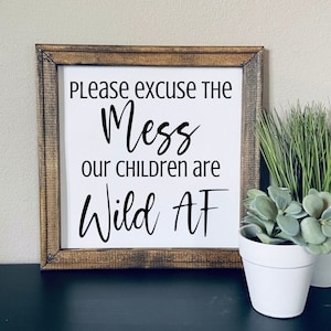 May include: A wooden framed sign with white background and black text that reads "Please excuse the mess our children are wild AF". The sign is sitting on a table with a potted plant in front of it.