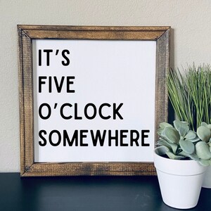 May include: A wooden framed sign with white background and black text that reads "It's Five O'Clock Somewhere".